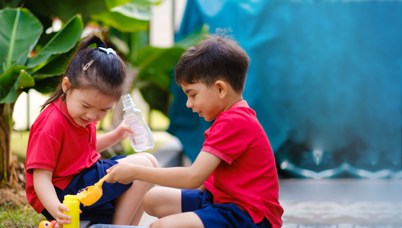 Two children in red Camberley uniforms enjoying water play in a leafy outdoor area